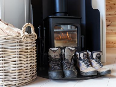 A pair of hiking boots next to a lit wood stove and a basket of firewood at High Longlands in Cartmel