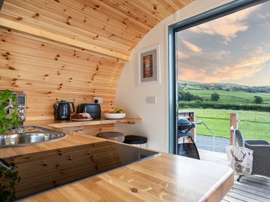 A kitchen with wooden counters and ceiling with tea kettle toaster and fruit bowl next to an open door showing a green landscape at High Longlands in Cartmel