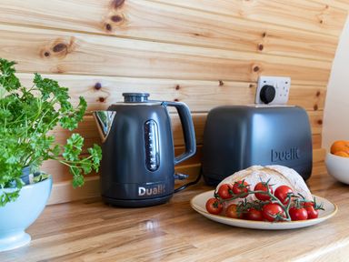 A kitchen counter with a black kettle, a black toaster, a plate with bread and cherry tomatoes, and a potted parsley plant at High Longlands in Cartmel