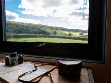 A table with a vintage camera, a camera case, and a map near a window showing a countryside view at High Longlands in Cartmel
