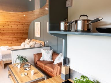 A kitchen area with pots on a shelf and a knife block overlooking a living area with a leather sofa and a bed at High Longlands in Cartmel