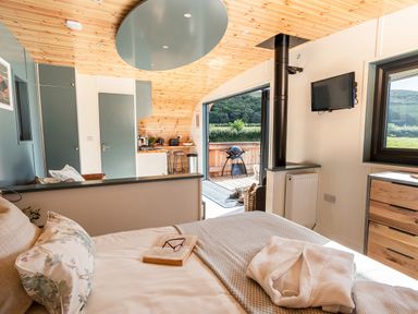 A bedroom with a bed a book glasses a TV mounted on the wall a window a kitchen area and an outdoor patio at High Longlands in Cartmel