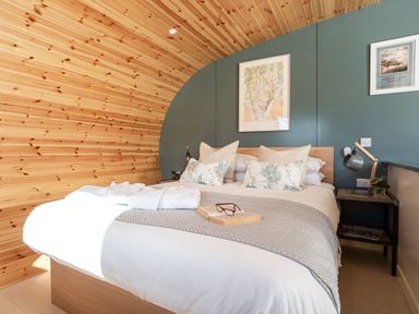 A bedroom with a wooden curved wall a bed with pillows and a throw and framed pictures on a teal wall at High Longlands in Cartmel