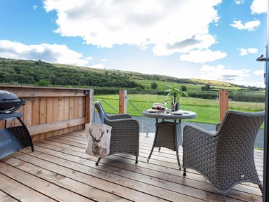 A wooden deck with two wicker chairs and a table set with wine glasses and a plant overlooking green hills at High Longlands in Cartmel