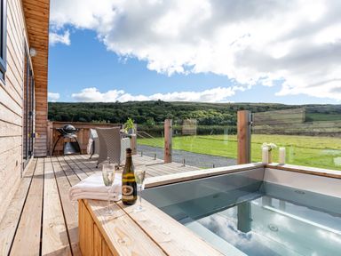 A wooden deck with a hot tub a bottle and two glasses a table and chairs and a barbecue overlooking green fields and hills at High Longlands in Cartmel