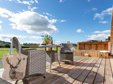 An outdoor wooden deck with wicker chairs a table with a plant bottle and glass a tote bag with a deer print and a wooden bench at High Longlands in Cartmel