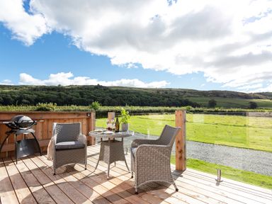 A wooden deck with two wicker chairs a round table with drinks and food and a barbecue grill with a grassy field and hills in the background at High Longlands in Cartmel