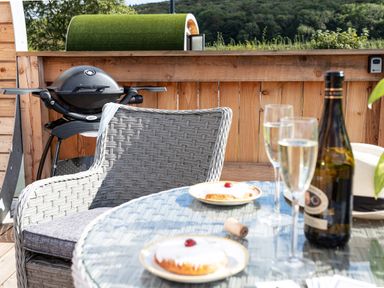 An outdoor patio with a wicker chair a grill a glass table with a bottle two glasses and two plates with pastries High Longlands Cartmel