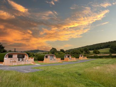 A row of four wooden cabins with green curved roofs on a grassy field under a sky with orange clouds at High Longlands in Cartmel