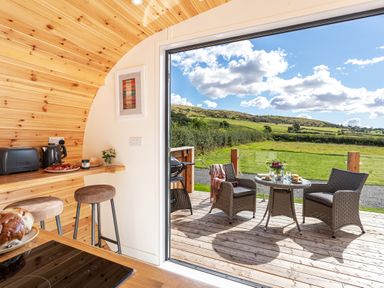 A small kitchen area with stools and appliances opening to a wooden deck with a table and chairs and a green landscape at Little Heaning in Cartmel
