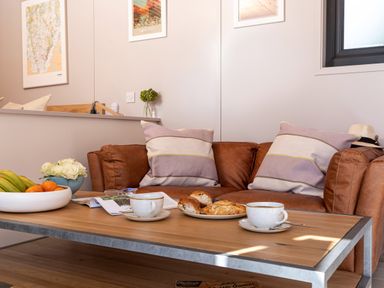 A living area with a brown leather couch and pillows with a wooden coffee table holding fruit bowls cups and pastries at Little Heaning in Cartmel