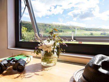 A wooden table with a vase of flowers binoculars a book and a hat by an open window showing a hill outside at Little Heaning in Cartmel