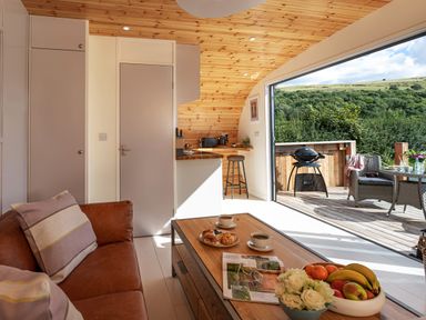 A living area with a brown sofa and wooden coffee table next to an open door leading to a patio with outdoor seating and a grill at Little Heaning in Cartmel