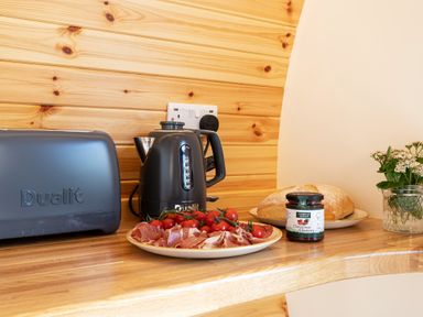 A kitchen counter with a toaster, electric kettle, plate of tomatoes and sliced meat, bread, jar of chutney, and a small plant at Little Heaning in Cartmel