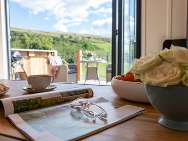 A table with a magazine holding glasses a cup a bowl of fruit and a bowl of white roses near an open door showing an outdoor patio with chairs and a table at Little Heaning in Cartmel