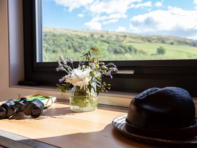 A table with a vase of flowers binoculars and a hat in front of a window showing a green hill outside at Little Heaning in Cartmel