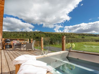 An outdoor wooden deck with a hot tub towels table chairs barbecue and grassy hills in the background at Little Heaning in Cartmel
