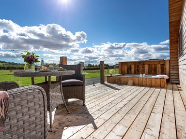 An outdoor wooden deck with wicker chairs a table with flowers and food and a wooden hot tub at Little Heaning in Cartmel