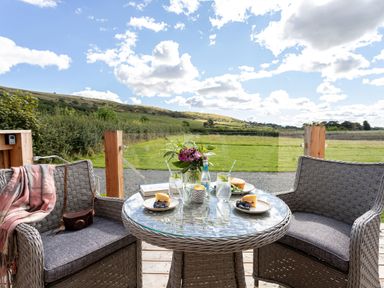 An outdoor wicker table with drinks and desserts and two wicker chairs on a deck overlooking fields at Little Heaning in Cartmel