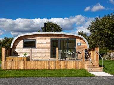 A wooden pod with a curved roof and veranda with chairs and a table at Little Heaning in Cartmel