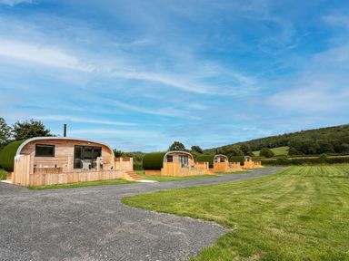 Four modern wooden glamping pods with green roofs on a grassy area with a gravel path and hills in the background at Little Heaning in Cartmel