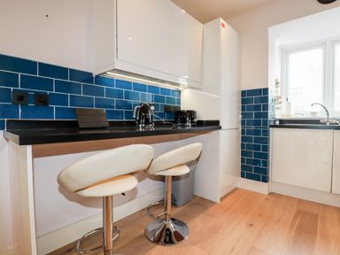 A kitchen with blue tiled backsplash black countertop two white bar stools and window at 6 Ocean Heights in Newquay