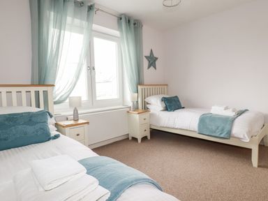 A bedroom with two single beds with blue cushions and throws by a window with blue curtains at 6 Ocean Heights in Newquay