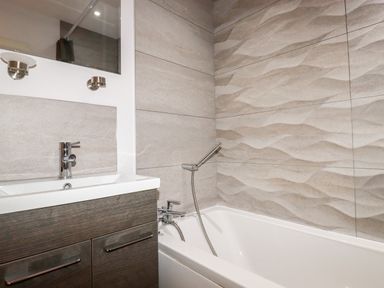 A bathroom with a bathtub a handheld shower a sink with a faucet a mirror and textured wall tiles at 6 Ocean Heights in Newquay