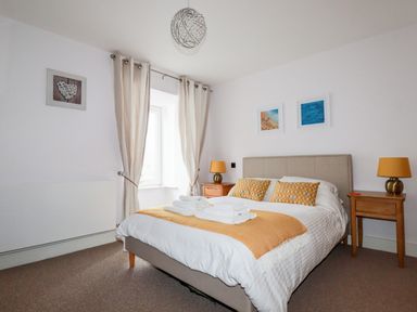 A bedroom with a bed with yellow pillows and a throw blanket two wooden bedside tables with lamps and framed pictures on the wall at 6 Ocean Heights in Newquay