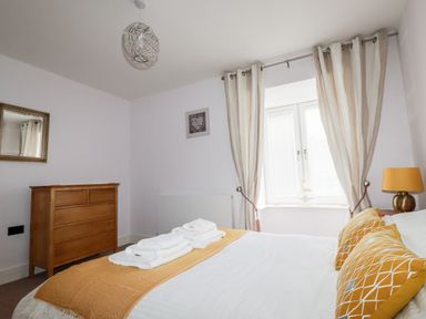 A bedroom with a bed with folded towels and mustard cushions a wooden dresser and a window with curtains at 6 Ocean Heights in Newquay