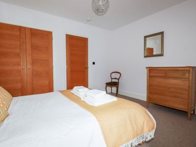 A bedroom with a bed covered with a beige blanket and folded towels a wooden dresser a chair two wooden doors and a mirror on the wall at 6 Ocean Heights in Newquay