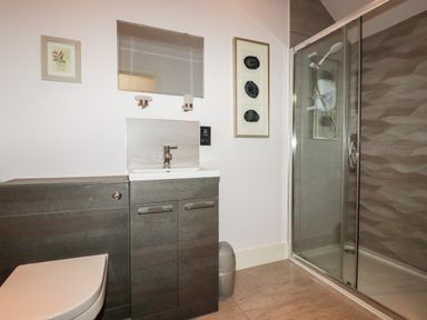A bathroom with a toilet, sink with cabinet, wall mirror, wall lights, framed pictures, and a glass shower enclosure at 6 Ocean Heights in Newquay