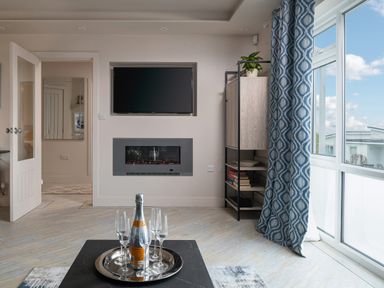 A living room with a wall-mounted TV and fireplace shelves with books and a plant and a bottle with glasses on a table near window with blue patterned curtains at Cartref in Benllech