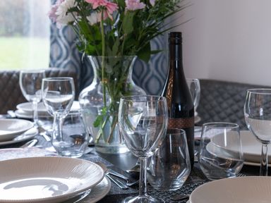 A dining table set with plates glasses cutlery a wine bottle and a vase with flowers at Cartref in Benllech