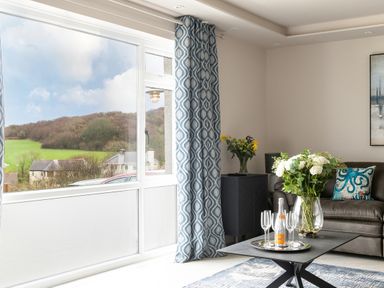 A living room with a leather sofa a coffee table with flowers and champagne and large window with blue curtains at Cartref in Benllech