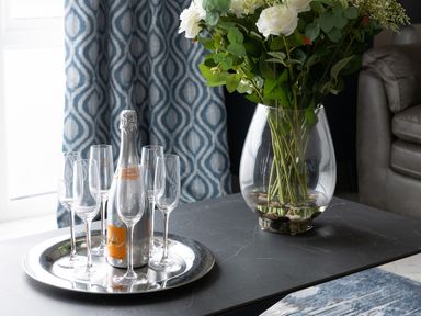 A table with a bottle of sparkling wine and six glasses on a tray and a vase with white flowers and green leaves at Cartref in Benllech