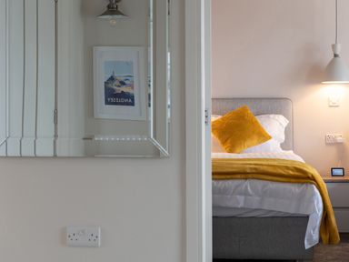 A bedroom with a bed with yellow pillow and blanket a bedside table a lamp and a mirror reflecting a framed picture at Cartref in Benllech