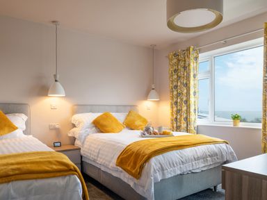 A bedroom with two beds yellow cushions and blankets a window with yellow curtains and a sea view at Cartref in Benllech