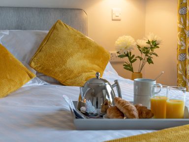 A bed with yellow cushions and a tray holding croissants juice and a teapot with flowers in the background at Cartref in Benllech