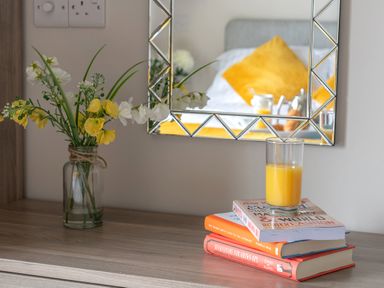A wooden table with a glass of orange juice on top of stacked books and a vase with yellow and white flowers in front of a wall mirror in a bedroom at Cartref in Benllech