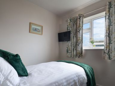 A bedroom with a single bed with green cushions and throw a wall-mounted TV and a window with floral curtains at Cartref in Benllech