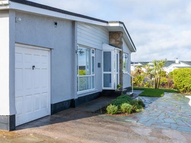 The exterior of a single-story house with a garage and a paved walkway surrounded by grass and plants at Cartref in Benllech