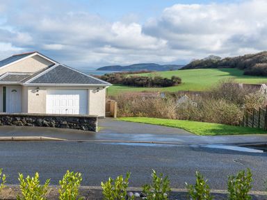 A house with a garage and driveway next to a paved road with green fields and hills in the background at Cartref in Benllech