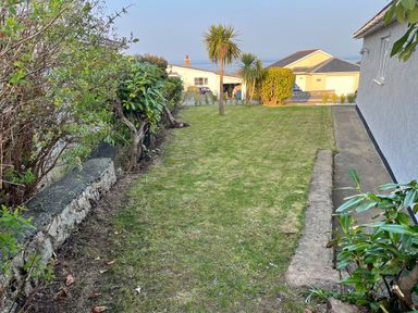 A grassy garden area with a concrete path along a house and various shrubs and palm trees at Cartref in Benllech