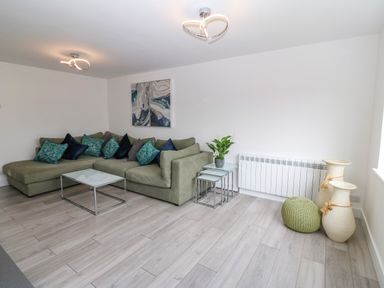 A living room with a green sectional sofa and blue cushions a glass coffee table and side tables with a plant and decorative vases at Daresfield in Cemaes Bay