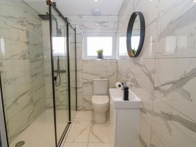 A bathroom with a glass shower enclosure a toilet a small sink with a black faucet a round mirror and a window with a plant on the sill at Daresfield in Cemaes Bay