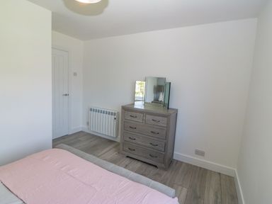 A bedroom with a bed covered in a pink blanket a wooden dresser with a mirror and a white door at Daresfield in Cemaes Bay