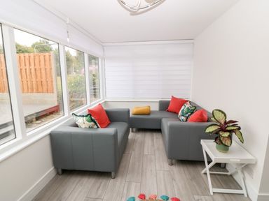 A seating area with gray sofas and colorful cushions by windows with white blinds and a white side table with a plant at Daresfield in Cemaes Bay