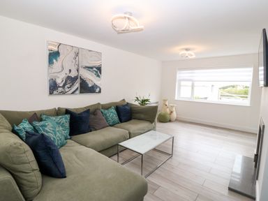A living room with a green sectional sofa with multiple cushions a glass coffee table wall art a large window with blinds and decorative vases at Daresfield in Cemaes Bay