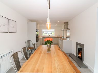 A dining area with a wooden table and metal chairs next to a kitchen with white cabinets and a fireplace at Daresfield in Cemaes Bay